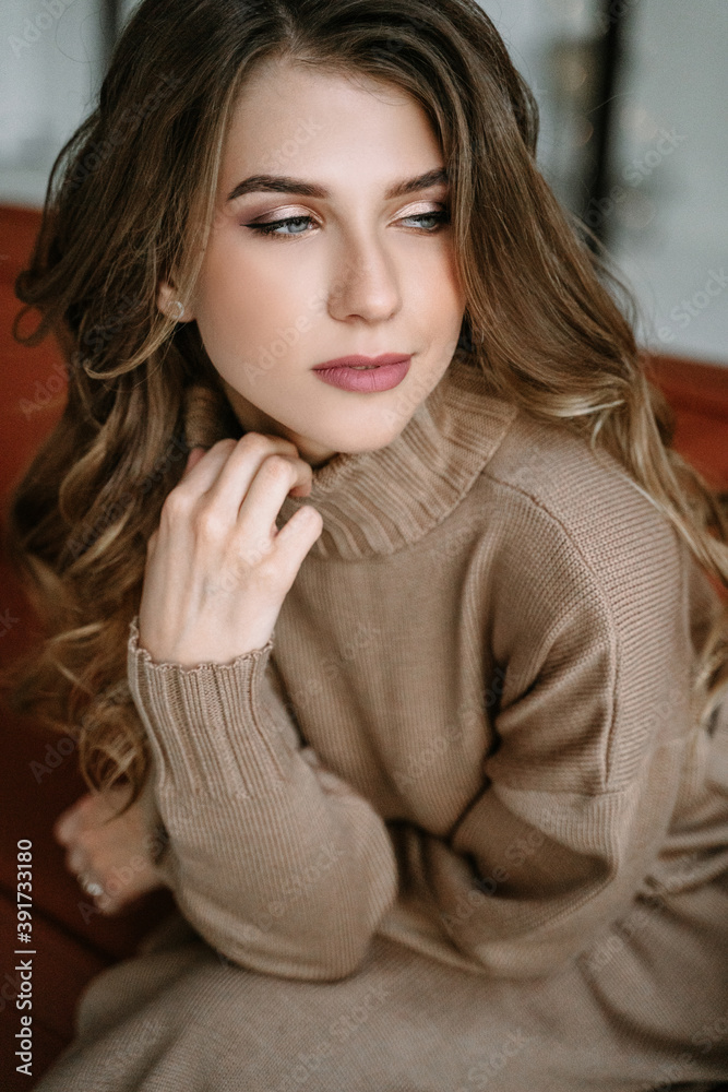 Blonde girl with long hair in a brown knitted dress in a stylish apartment