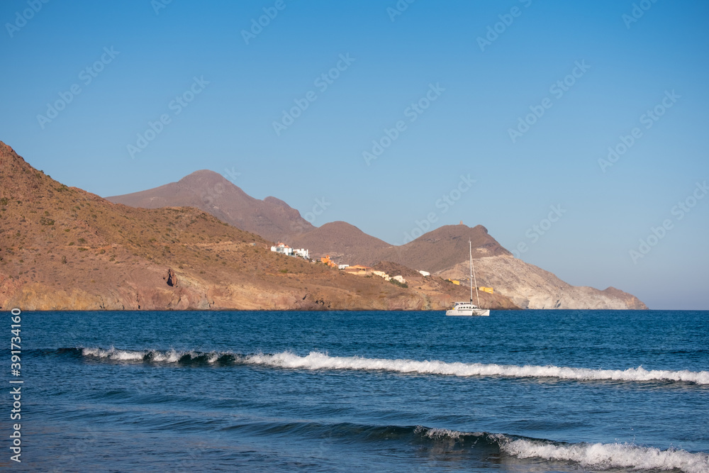 Recreational boat anchored in front of the beach, village with colorful houses on the hill, desert landscape by the Mediterranean sea. Playa de los genoveses, San José, Almería, Andalusia, Spain