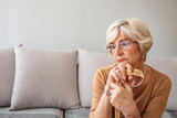 Senior woman sitting on sofa and holding walking stick. Alone disappointed upset 70s woman sitting on sofa hides her face with hands holding walking stick  suffers from physical disability disease