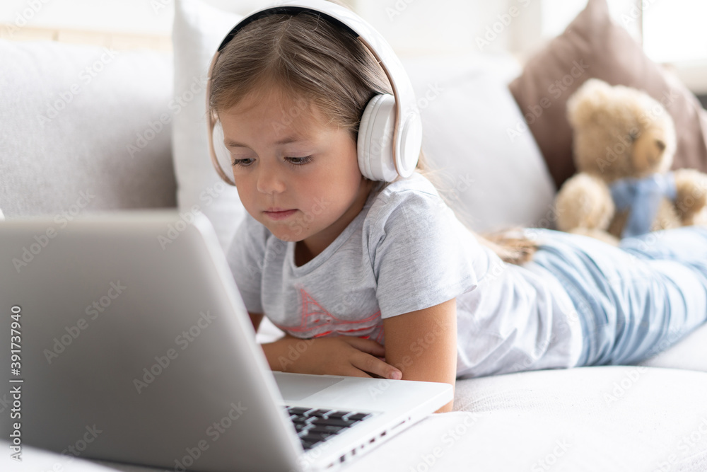 Online education of children. Sweet girl with headphones looking video lesson teacher conference laptop sitting on the couch at home. Online education of children. Sweet girl with headphones looking video lesson teacher conference laptop sitting on the couch at home.