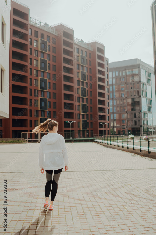 Naklejka premium A young blonde sports girl in a black suit runs along modern houses