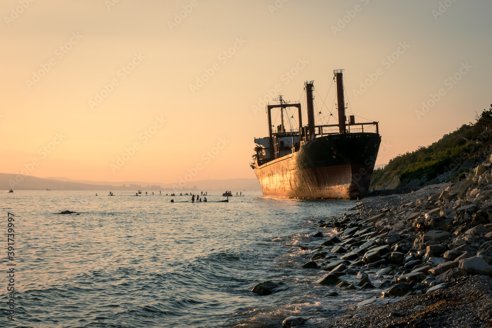 Big cargo ship running aground. Sunset on the pebbly seashore. Silhouettes of people on SUP boards on a sunset background