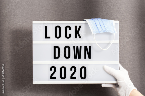 Hands in white gloves hold Light box with message lockdown 2020 and Surgical protective mask. Word of the Year 2020 is lockdown. photo