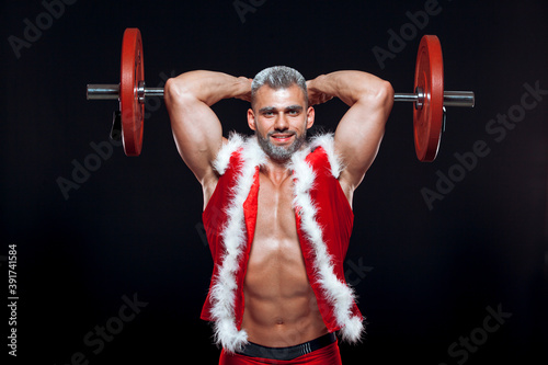 Sexy Santa Claus. Bodybuilder young handsome santa clause smile train muscles arms and triceps at New Years eve and Christmas winter holiday black background.