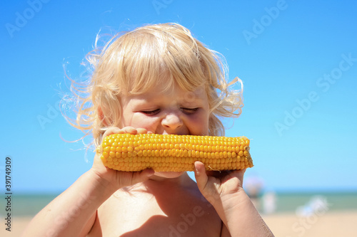 Little blond boy eating boiled corn on the beach. Healthy food, summertime, vacation concept.