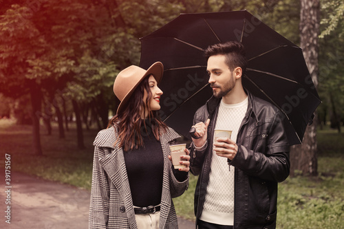 Young couple walking spring green city park, drinking hot coffee paper eco recycle cup. Two lovers husband wife spending time together, discuss news movie film with black umbrella to shelter the rain.