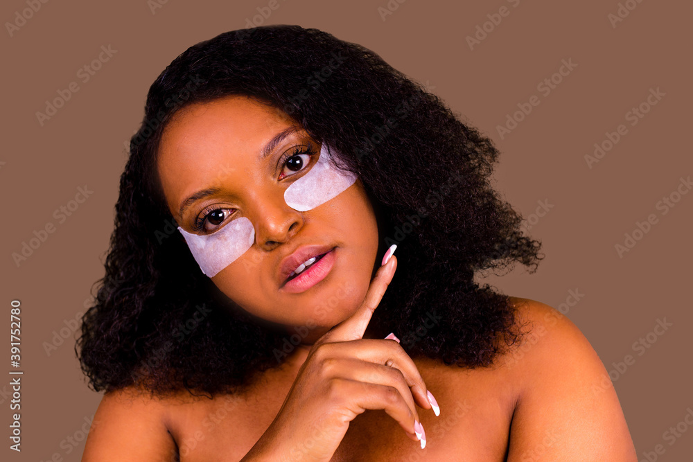 Fototapeta premium Attractive woman applying anti-fatigue under-eye mask in studio brown background