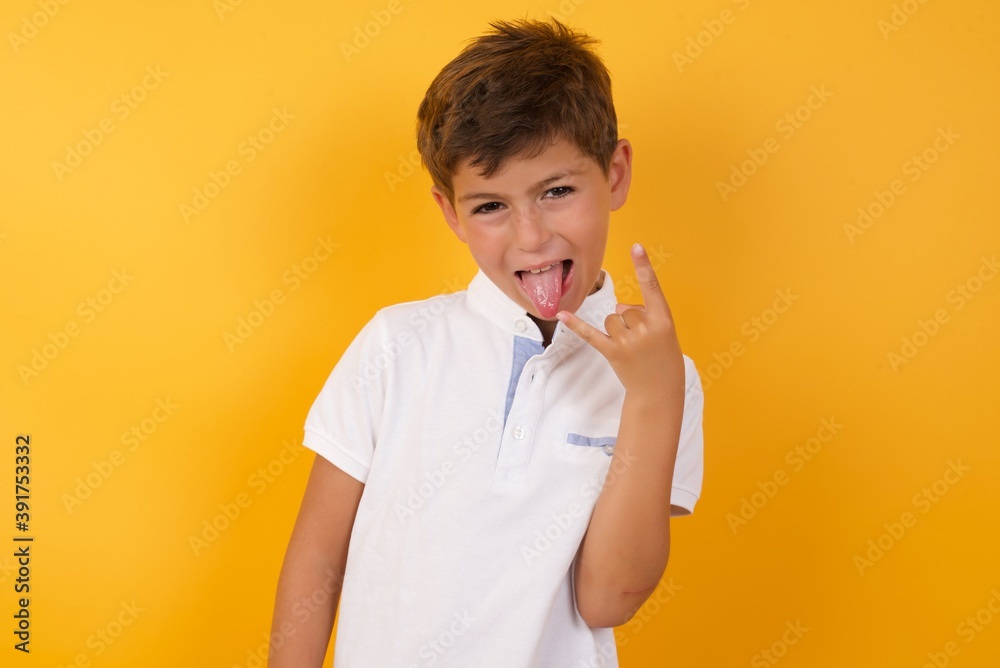 Portrait of a crazy Cute Caucasian little boy standing against yellow background showing tongue horns up gesture, expressing excitement of being on concert of band. Portrait of a crazy Cute Caucasian little boy standing against yellow background showing tongue horns up gesture, expressing excitement of being on concert of band.