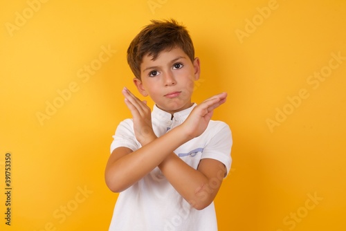 Cute Caucasian little boy standing against yellow background Has rejection angry expression on face and crossing hands doing refusal negative sign.