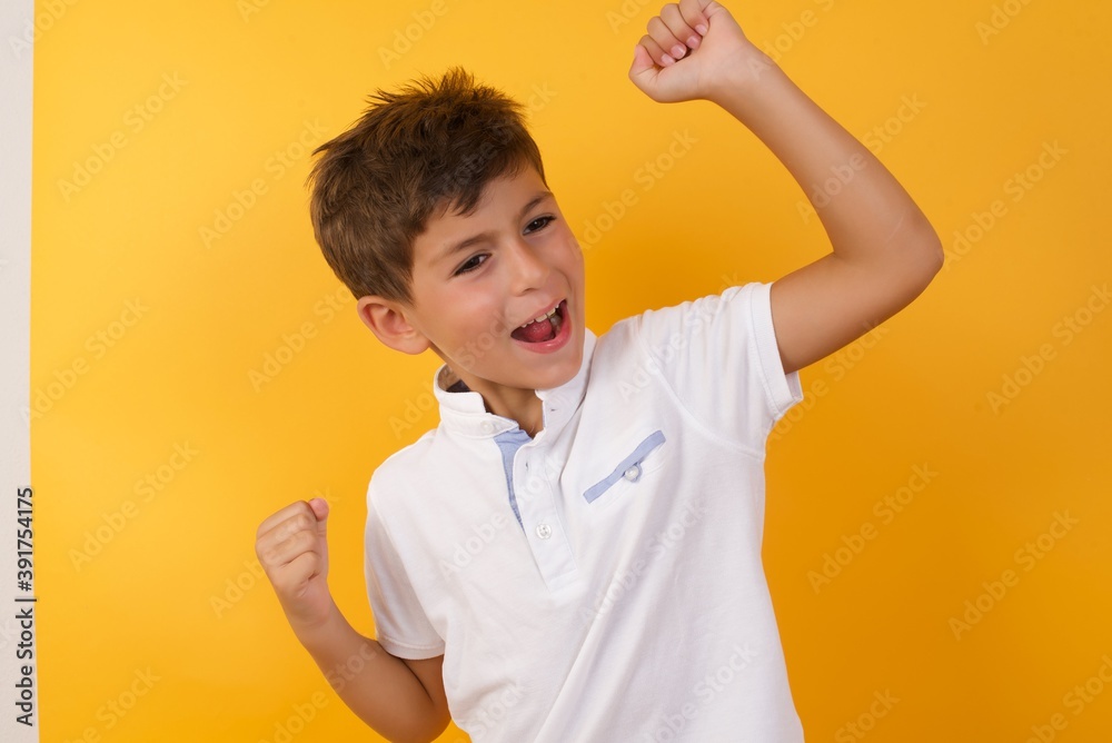 Attractive Young handsome Caucasian little boy standing against yellow wall celebrating a victory punching the air with his fists and a beaming toothy smile Attractive Young handsome Caucasian little boy standing against yellow wall celebrating a victory punching the air with his fists and a beaming toothy smile