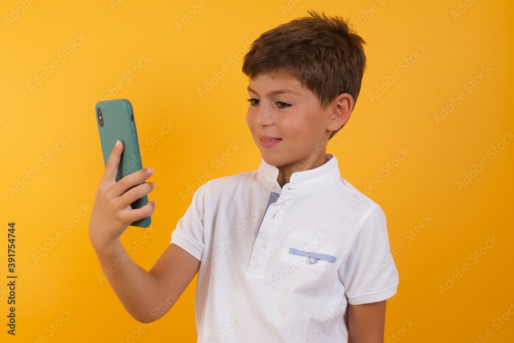 Isolated shot of pleased cheerful Young handsome Caucasian little boy standing against yellow background , makes selfie with mobile phone. People, technology and leisure concept Isolated shot of pleased cheerful Young handsome Caucasian little boy standing against yellow background , makes selfie with mobile phone. People, technology and leisure concept