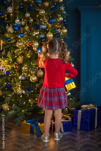 A cute girl in a red dress holds a gift box in her hands and touches the decorations on the Christmas tree, under which there are many gifts