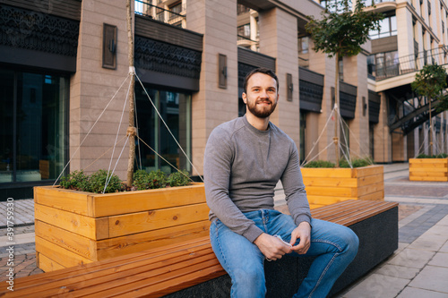 Cheerful young bearded man sitting on urban bench at modern European city street. Relaxed handsome male having resting outdoors on empty street. City on quarantine. Commercial business shutdown.