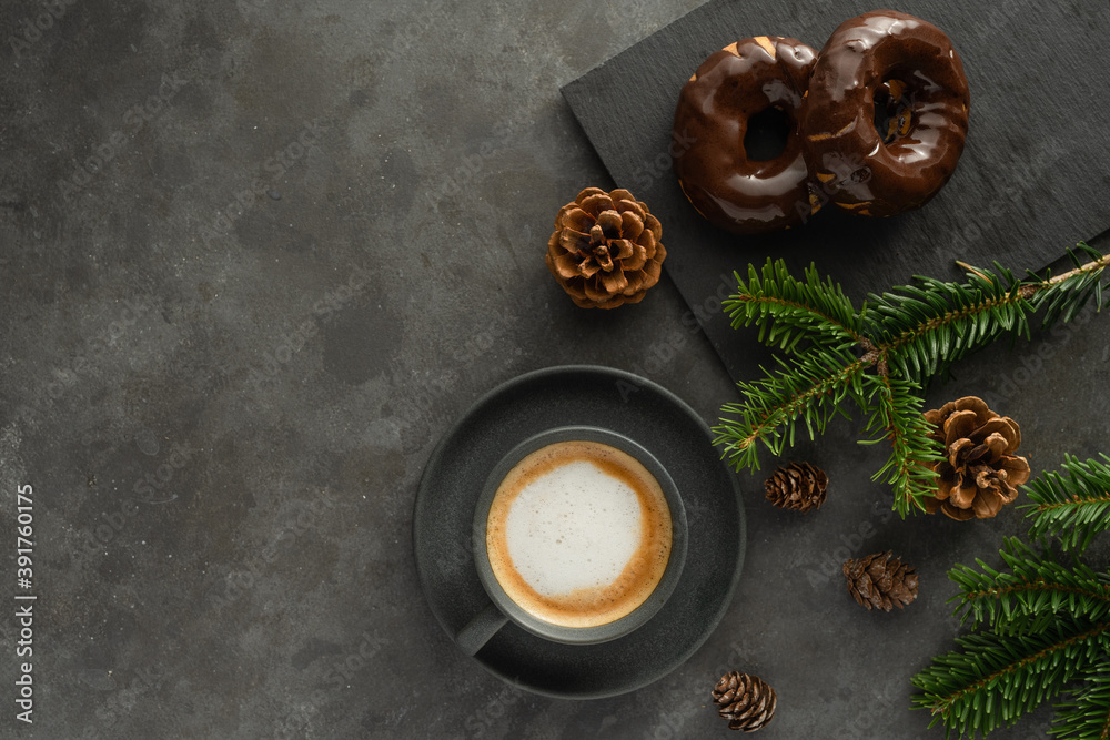 A photo of cappuccino with chocolate donuts and christmas decoration.  Grey cups on the grey table and a grey wall. Place for text