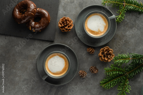 A photo of cappuccino with chocolate donuts and christmas decoration. Grey cups on the grey table and a grey wall. Place for text © Malin