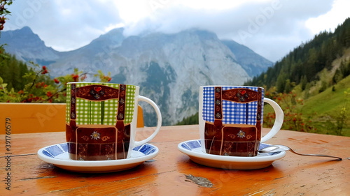 Two coffee mugs with traditional dirndl outfit painting on an outdoor table against the beautiful mountains of the Austrian Alps.