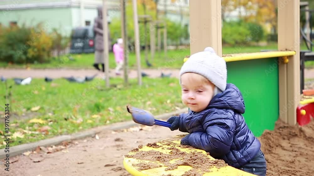 Funny little blond boy in a warm jacket and hat playing with plastic toys molds in the sandbox on the Playground in the Park. Happy childhood. Game Funny little blond boy in a warm jacket and hat playing with plastic toys molds in the sandbox on the Playground in the Park. Happy childhood. Game