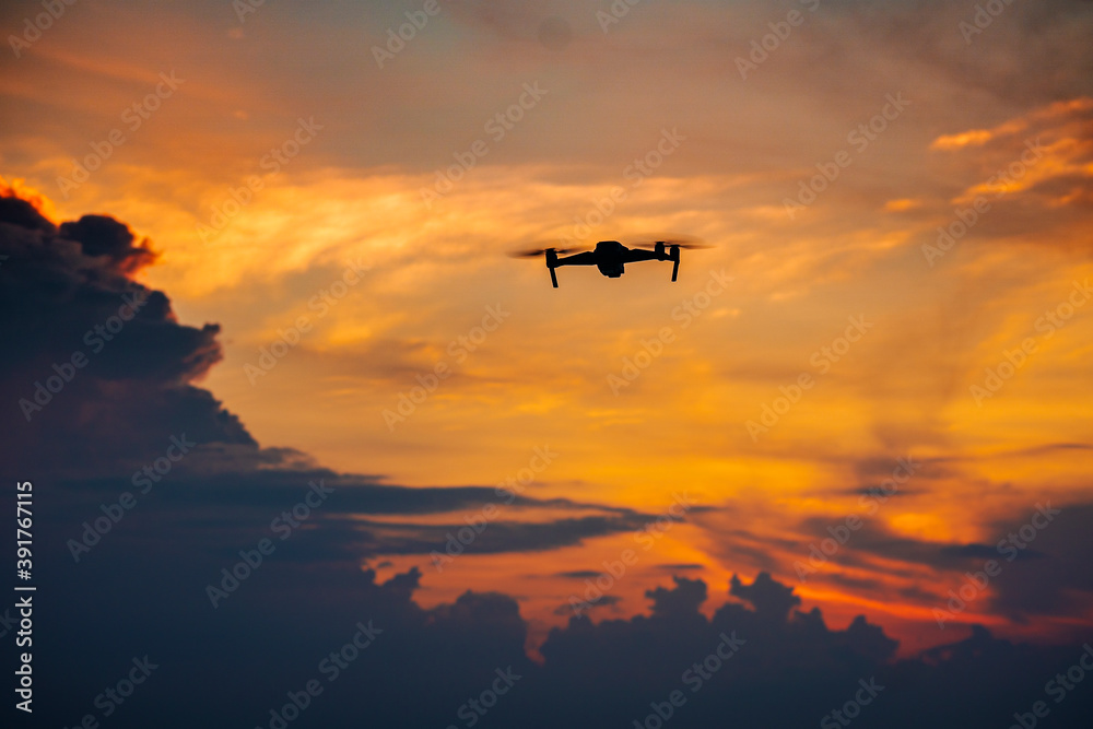 Modern technological camera concept. Silhouette of drone flying in glowing orange sunrise sky in the morning over nature background.