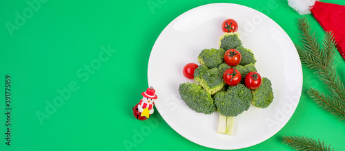 Christmas tree of organic vegetables on white plate; Broccolis and tomatoes with fork and knife on green background. Merry Xmas and happy New Year party, New You, Healthy food and vegetarian concept
