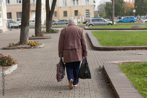 An old lonely elderly poor gray-haired woman with bags walks from the store or down the street. Rear view, face not visible.