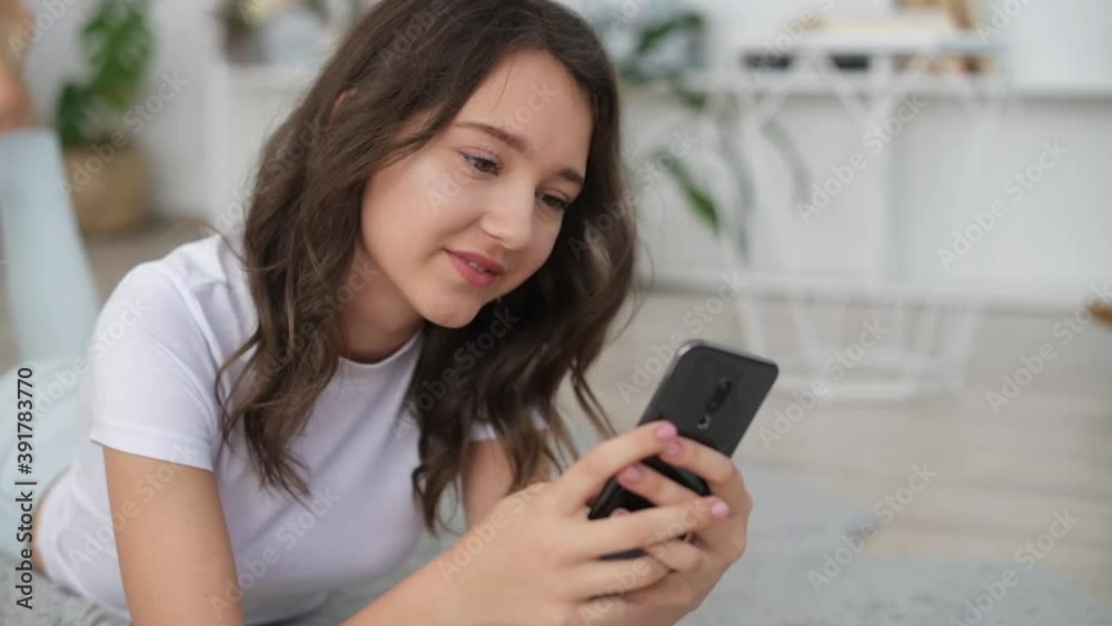 Young cheerful girl lying down on the ground indoors and using smartphone. Young cheerful girl lying down on the ground indoors and using smartphone.