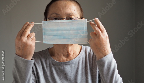 Portrait of an elderly woman wearing a protective medical mask, looking at the camera, on a gray background. Flu epidemic, dust allergy, virus protection. City air pollution concept.  photo