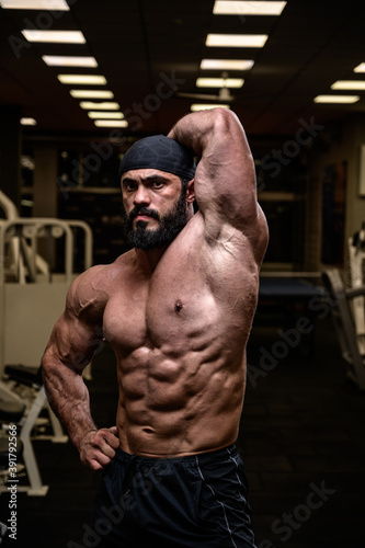 strong bearded male in bandana showing perfect sport physique with abdominal oblique muscle in dark fitness gym