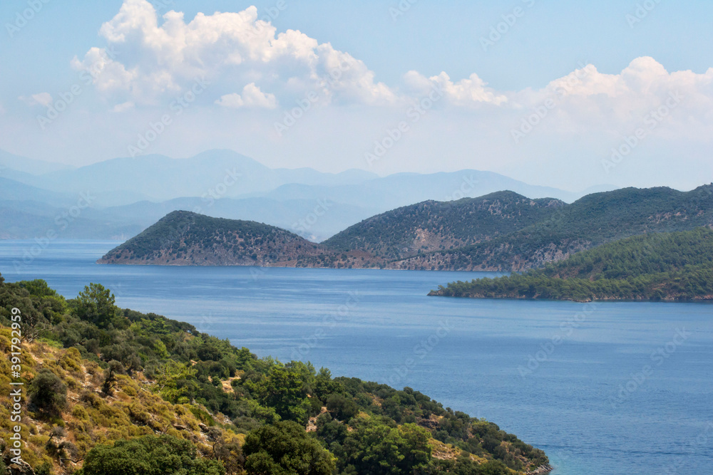 A bright blue ocean surrounded by hills covered with green foliage with clouds and bright blue skies in the background.