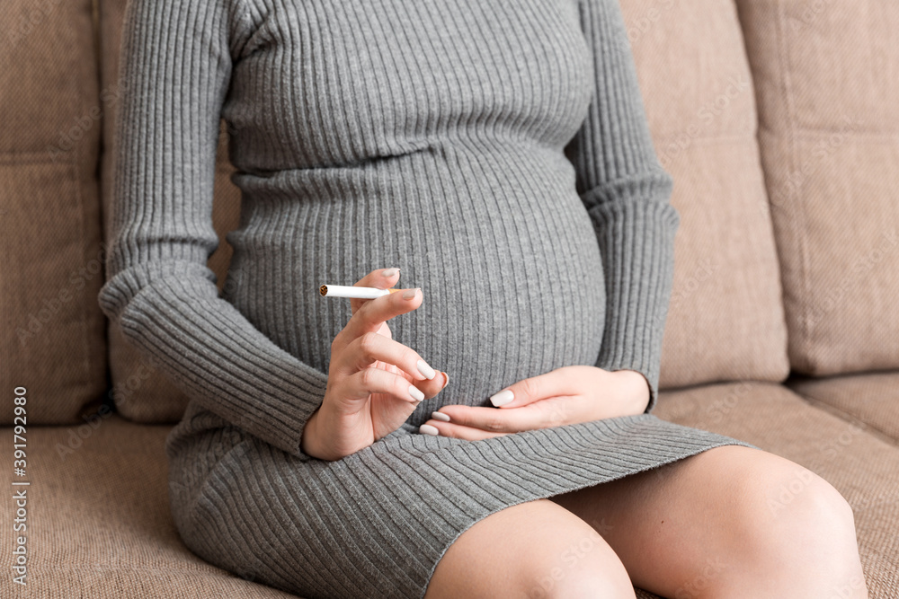 Close-up of pregnant woman is holding a cigarette sitting on sofa at home