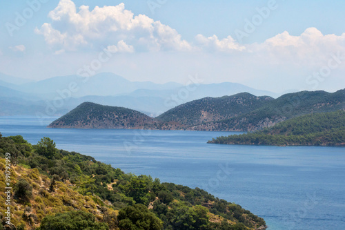A bright blue ocean surrounded by hills covered with green foliage with clouds and bright blue skies in the background.