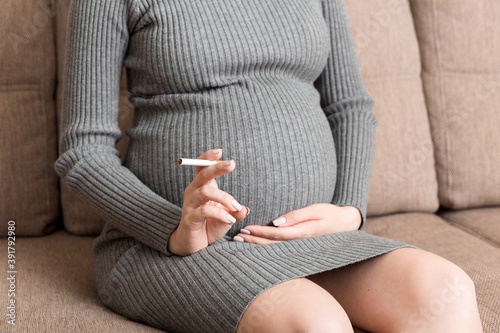 Close-up of pregnant woman is holding a cigarette sitting on sofa at home © sosiukin