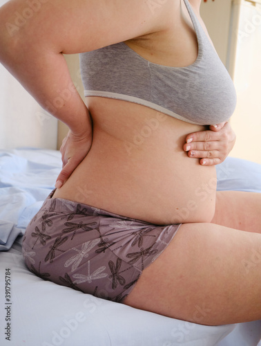 Back pain of a pregnant woman sitting on a bed in the bedroom, holding her lower back and stomach. Profile of the expectant mother's naked belly