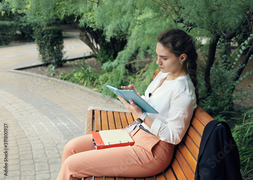 Young beautiful female student calmly reads in tablet while sitting on park bench alone