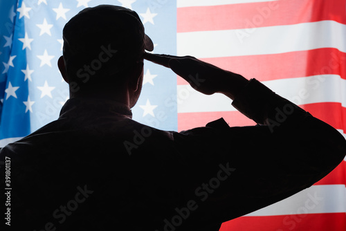 back view of patriotic military man in uniform and cap giving salute near american flag photo