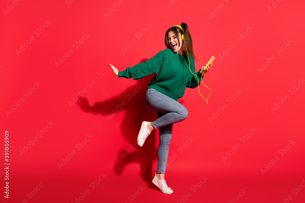 Full size photo of young happy crazy smiling girl hold mobile listen music in yellow earphones isolated on red color background