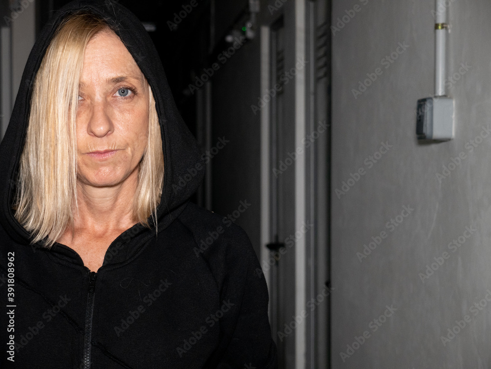 BLOND SPANISH WOMAN DRESSED IN BLACK HOODED COAT, IN THE CORRIDOR OF A BASEMENT WITH A SINIEST LOOK