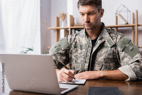 Canvas Print military man in uniform holding pen while writing on clipboard near laptop on de
