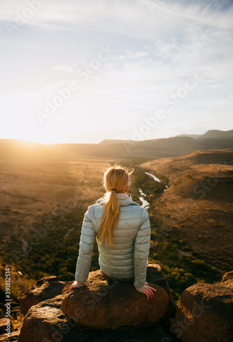 Free-spirited young teen sitting relaxing on boulder on top on luscious mountain watching sunset 