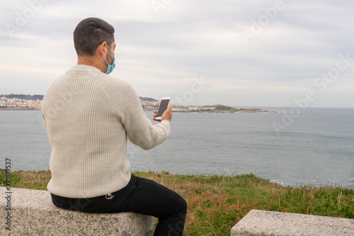 Man with mask sitting checking his cell phone in a beautiful viewpoint near the sea