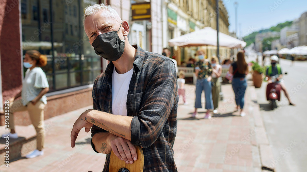 Portrait of middle aged man wearing mask holding a longboard and looking at camera. People collecting their orders from the pickup point during coronavirus lockdown in the background Portrait of middle aged man wearing mask holding a longboard and looking at camera. People collecting their orders from the pickup point during coronavirus lockdown in the background