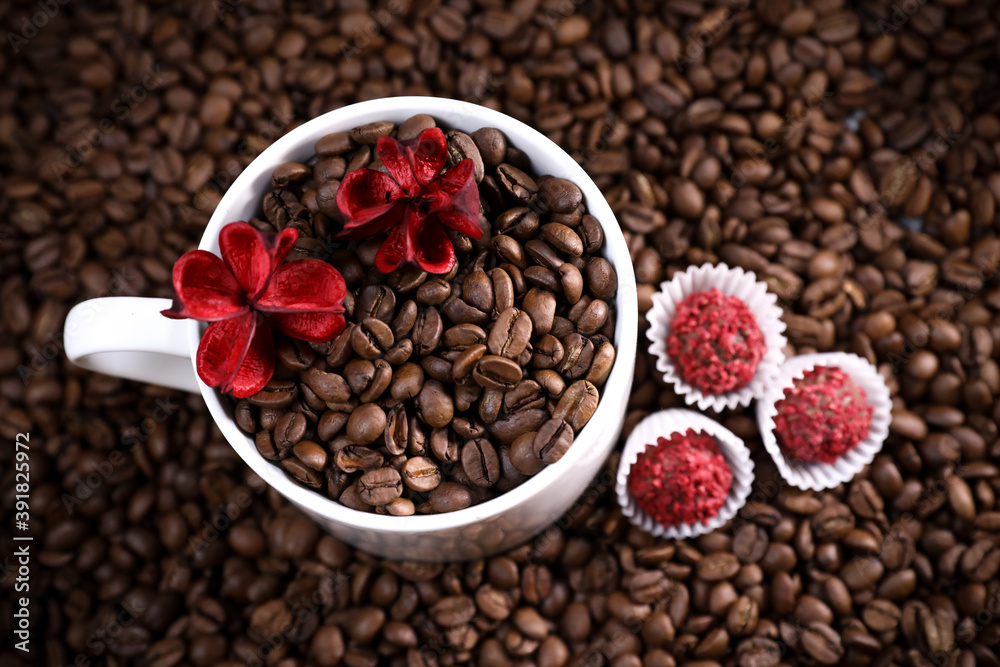 Fototapeta premium White cup full of coffee beans on a coffee beans background with red chocolate candy and red flowers. Morning espresso. Coffee mug. 