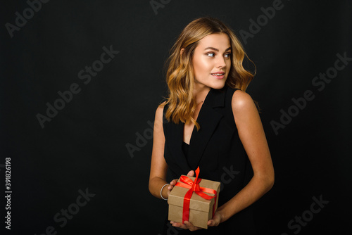 A beautiful young woman on a black background holding a Christmas present with a red ribbon. The happiness of receiving and giving gifts for the new year