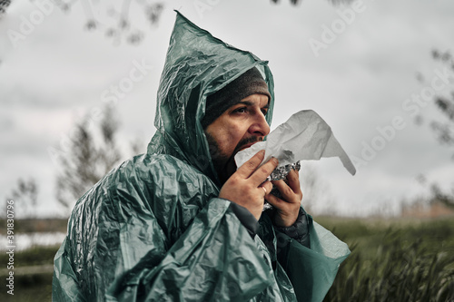 A man with a beard in a green raincoat eats in nature. Cloudy weather, fast food.