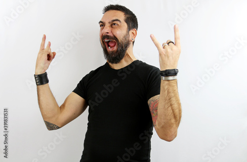 A young man, with a beard, black t-shirt and tattoos on a white isolated background, yells with crazy expression doing the sibble of rock with his hands up. Heavy metal concept