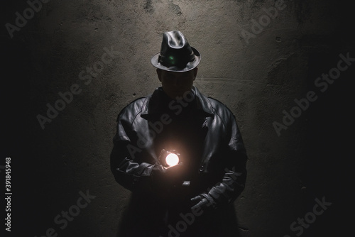 Detective agent in black coat and in leather hat is standing near the building wall at night.