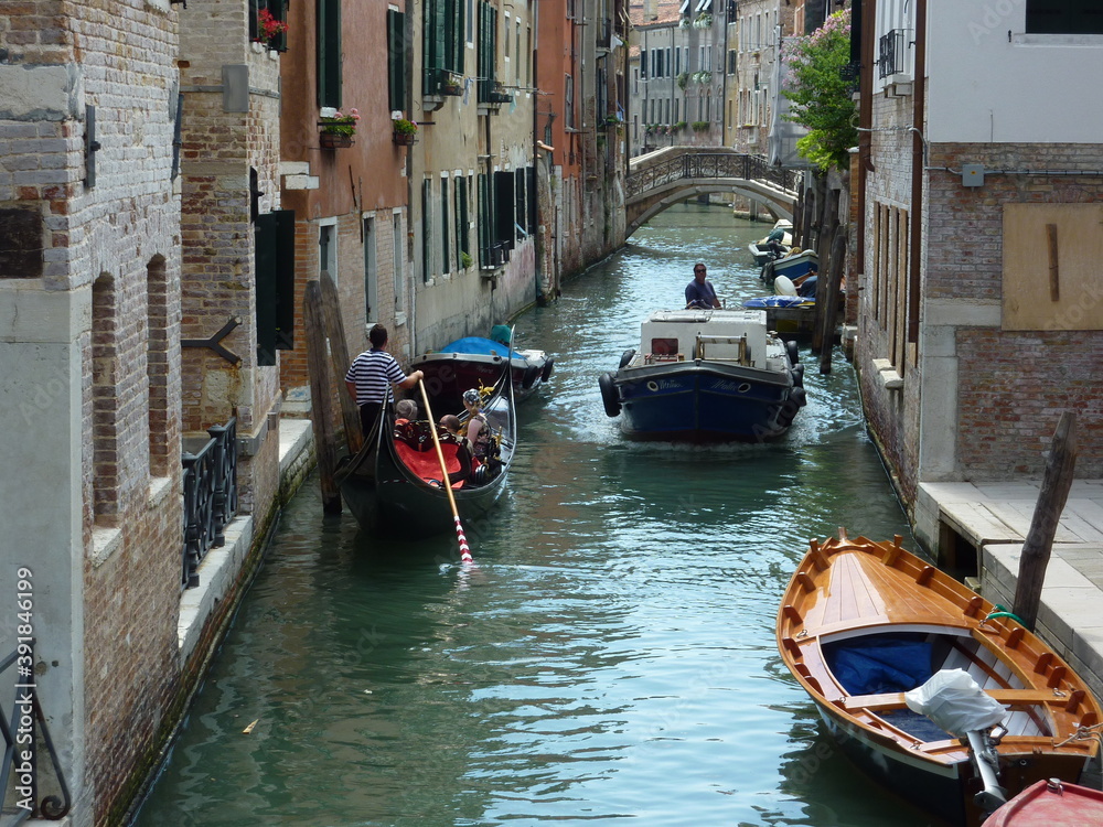 A gondola gives way to a motorboat in a narrow Venetian canal. A bridge over the canal and boats moored to its banks.