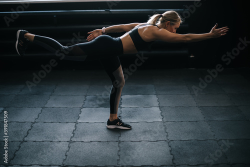 Fitness young woman with perfect athletic body wearing black sportswear doing warrior three yoga pose standing on one leg leaning forward with chest and leg parallel to the floor working out in gym.