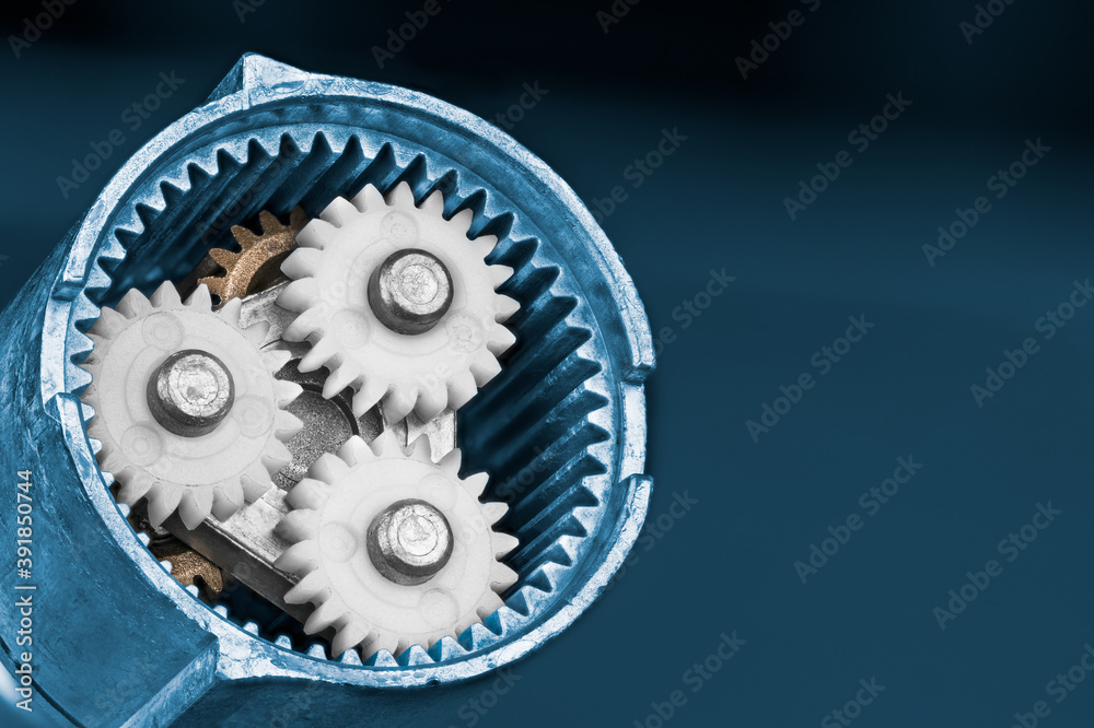 White planet gears inside a ring cogwheel of epicyclic gear train on blue background. Close-up of plastic and metallic cog wheels in disassembled gearbox of electric screw gun. Mechanical engineering. White planet gears inside a ring cogwheel of epicyclic gear train on blue background. Close-up of plastic and metallic cog wheels in disassembled gearbox of electric screw gun. Mechanical engineering.