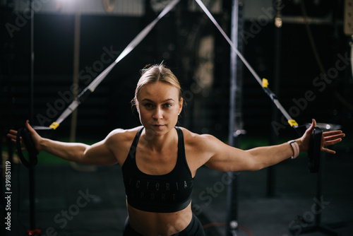 Middle shot portrait of sporty young woman with perfect muscular body wearing black sportswear working out on simulator during sport training at fitness gym with dark interior, looking at camera.