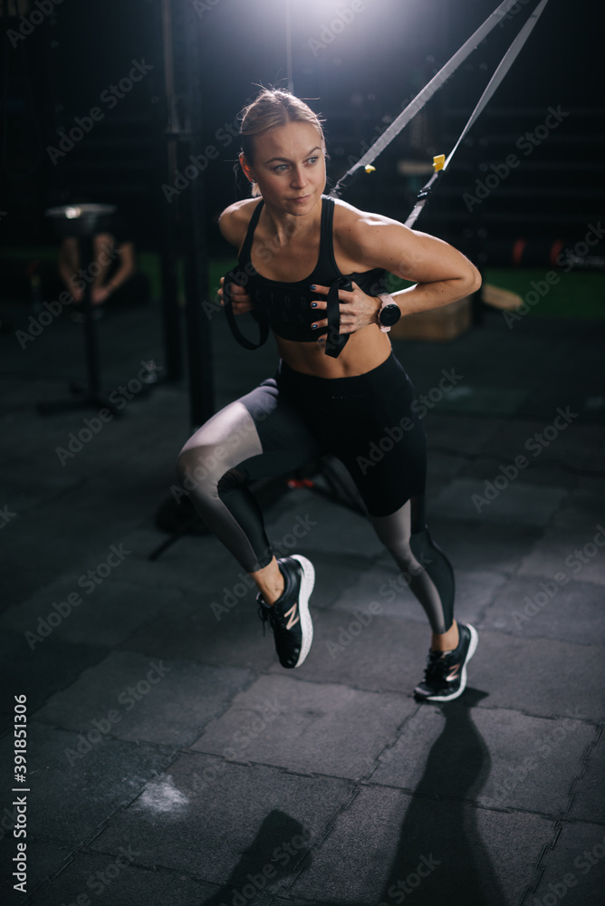 Sporty young woman with perfect muscular body wearing black sportswear working out on simulator during sport training at modern fitness gym with dark interior.
